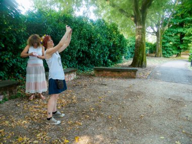 Açık havada güneşli bir günün tadını çıkaran iki genç kadın, yemyeşil ağaçlar ve doğal ışık altında çekilmiş yapraklarla dolu bir parkta fotoğraf çektirmek için poz veriyor.