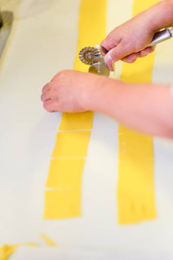 Temiz beyaz bir yüzey üzerinde tırtıklı bir tekerlek makasıyla uzun sarı erişteleri keserek taze makarna hamuru hazırlamak mutfak sanatı ve yemek fotoğrafçılığı için lezzetli İtalyan mutfağı yaratmak.