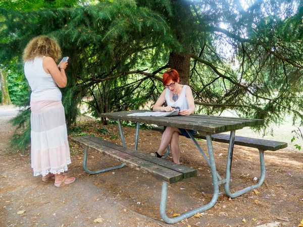 İki kadın, doğa ve yapraklarla çevrili ağaçların gölgesinde piknik masasında huzurlu bir öğleden sonranın tadını çıkarıyor. Bir bağ ve rahatlama anı yakalıyorlar.