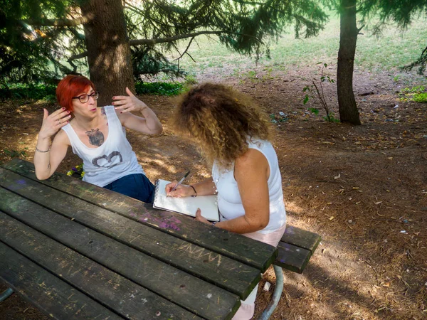 Güneşli bir günde parkta piknik masasında sohbet eden iki kadın arkadaş.