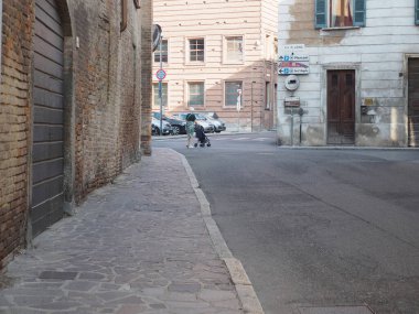 Cremona, İtalya 6 Ağustos 2025 Parlak yaz öğleden sonra İtalya 'da tarihi binalardan ve park halindeki araçlardan geçen bir kadın bebek arabasını kaldırım taşı caddesinde itiyor.