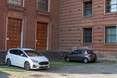 Cremona, İtalya - 2 Ekim 2025 Büyüleyici ahşap kapıları olan geleneksel tuğla mimarinin önünde beyaz bir Ford S-Max ve gri bir Toyota Yaris bir kaldırım taşı parkına park edilmiştir.