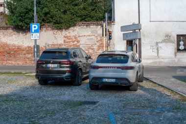 Cremona, İtalya - 2 Ekim 2025 Premium lüks SUV 'ler (BMW X5, Porsche Macan) tarihi binalar ve tabelaların yanında güneşli bir Avrupa kaldırım taşı caddesine park ettiler.