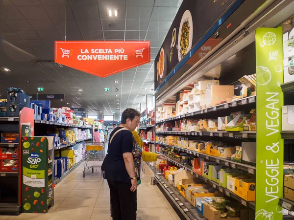 Cremona, İtalya - 17 Eylül 2025 Bir kadın Kuzey İtalya 'daki bir süpermarket koridorunda çeşitli ürünlerle dolu raflardan dikkatlice bakkaliye seçti