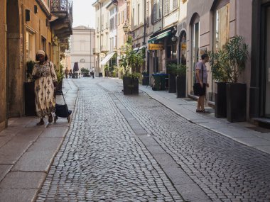 Cremona, İtalya 13 Ağustos 2025 Afrikalı bir kadın, güneşli bir yaz gününde, İtalya 'nın Cremona şehrinde bir alışveriş arabasıyla bir kaldırım taşı caddesinde yürüyor.