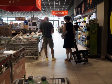 Cremona, İtalya - 15 Ağustos 2025 Bir çift tüketici, çeşitli mağaza bölümleri ve ürün kategorilerini gösteren sayısız alışveriş arabaları ve tepe tabelaları bulunan parlak bir süpermarket koridorunda ürünleri denetliyor
