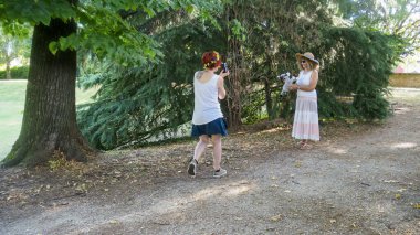Açık havada güneşli bir günün tadını çıkaran iki kadın, yemyeşil ağaçlar ve benekli güneş ışığıyla çevrili bir parkta poz vererek rahat bir yaşam tarzı portresi için huzurlu bir ortam yaratıyor.