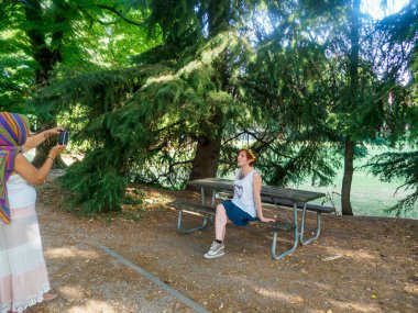 Parkta güneşli bir günün tadını çıkaran iki kadın arkadaş piknik masasının yanında dalların arasından süzülen benekli güneş ışığıyla poz veriyor.