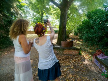 Parkta güneşli bir günün tadını çıkaran iki kadın bir kadının kafasında çiçek tacıyla fotoğraf çekiyorlar ve ellerinde ağaçlar ve yere saçılmış yapraklarla bir kamera var.