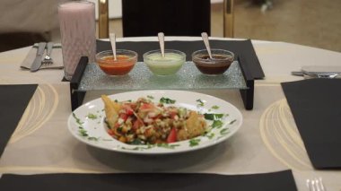 Geleneksel Hint samosa chaat beyaz bir tabakta servis edilir, yanında üç farklı sos ve zarif bir restoran masasına yerleştirilmiş bir bardak Lassi vardır.