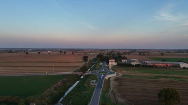 Po Valley fields stretching to the horizon at twilight, modern roundabout linking new roads beside an irrigation canal, showcasing sustainable agriculture and rural infrastructure development