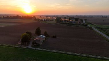 Hava görüntüsü, gün batımında tarımsal tarlalardan ve tarım arazilerinden geçen küçük bir arabayı yakalıyor. Ilık tarafından aydınlatılmış. Piacenza yakınlarındaki İtalyan kırsalında canlı turuncu gökyüzü