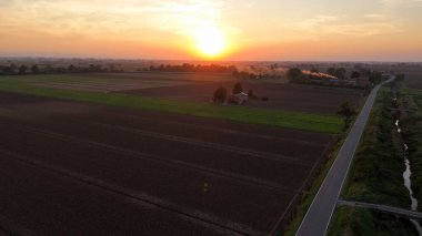 Hava görüntüsü, gün batımında tarımsal tarlalardan ve tarım arazilerinden geçen küçük bir arabayı yakalıyor. Ilık tarafından aydınlatılmış. Piacenza yakınlarındaki İtalyan kırsalında canlı turuncu gökyüzü