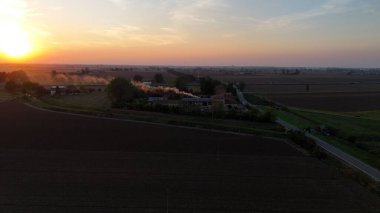 Hava görüntüsü, gün batımında tarımsal tarlalardan ve tarım arazilerinden geçen küçük bir arabayı yakalıyor. Ilık tarafından aydınlatılmış. Piacenza yakınlarındaki İtalyan kırsalında canlı turuncu gökyüzü