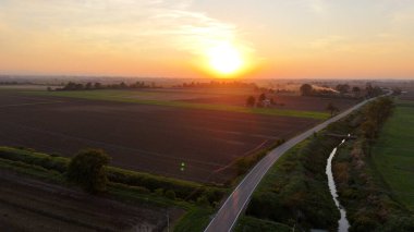 Hava görüntüsü, gün batımında tarımsal tarlalardan ve tarım arazilerinden geçen küçük bir arabayı yakalıyor. Ilık tarafından aydınlatılmış. Piacenza yakınlarındaki İtalyan kırsalında canlı turuncu gökyüzü