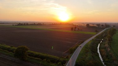 Hava görüntüsü, gün batımında tarımsal tarlalardan ve tarım arazilerinden geçen küçük bir arabayı yakalıyor. Ilık tarafından aydınlatılmış. Piacenza yakınlarındaki İtalyan kırsalında canlı turuncu gökyüzü
