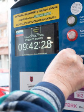 Sezana, Slovenya - 22 Kasım 2025 Hand, Slovenya 'nın Sezana kentinde bulunan dijital ekran görüntüleme süresi, bir Slovenya bayrağı ve ödeme talimatlarının yer aldığı modern bir parkmetreyle etkileşime girdi