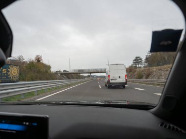 Sezana, Slovenya - 22 Kasım 2025 Sürücü perspektifi bulutlu bir günde hareket eden bir arabanın içinden boş bir otoyolda ilerleyen beyaz bir minibüsü yakalayarak İtalya 'ya yaklaşıyor.