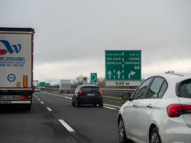Friuli, İtalya 21 Kasım 2025 'te Milano, Venezia, Udine ve Tarvisio' yu gösteren yeşil İtalyan A4 otoyol tabelası yoğun bir otoyolda bulutlu bir gökyüzünün altında