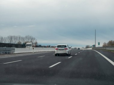 Friuli, İtalya 21 Kasım 2025 Beyaz araba bulutlu bir gökyüzü altında modern bir İtalyan otoyolu boyunca seyahat, hareket ve temiz yol altyapısını vurguluyor
