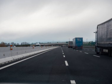 Friuli, İtalya 21 Kasım 2025 Kırsal karayolu birden fazla şeritli, kamyonlu ve otomobilli sağlam beton güvenlik bariyerlerini geçerek bir üst geçidin altından geçen, Tramvay 'ın üzerinde bulutlu bir gökyüzü görünüyor.