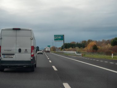 Venedik, İtalya 21 Kasım 2025. Beyaz minibüs bulutlu gökyüzünün altında otoyol boyunca trafik, yol işaretleri ve sonbahar ağaçlarıyla birlikte A4 otoyolu boyunca seyahat ediyor.