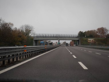 Venedik, İtalya 21 Kasım 2025 İtalya 'nın A4 otoyolunda ilerleyen ağır kamyon şeritleri, bulutlu gri bir günde modern bir üstgeçidin altından geçiyor.