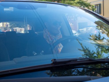 Kadın şehir caddesinde araba sürüyor, ön camdan yansımaları görünüyor.