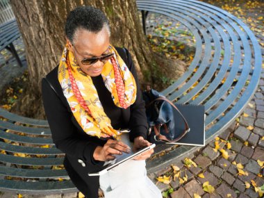 Parktaki bankta oturan yaşlı bir kadın, dışarıda dijital tabletle meşgul.