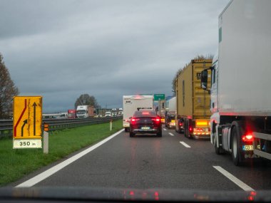 21 Kasım 2025 'te İtalya' nın Brescia kentinde, ıslak A21 otoyolunda trafik, sağanak yağış nedeniyle duraksamakta, arabalar ve kamyonlar bulutlu bir gökyüzü altında yavaşça hareket etmektedir. Şerit birleştirme işareti,