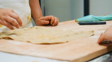 Çocuğun elleri ahşap bir yüzeyde taze bir hamur parçasını yoğurup şekillendiriyor, pişirme sanatını öğreniyor ve mutfak ortamında geleneksel hamur işleri yapıyor.