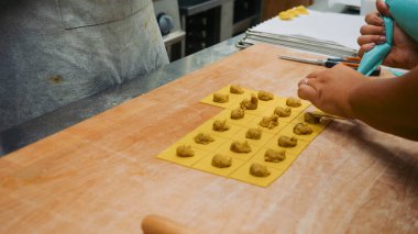 Şefin elleri özenle taze makarna hamurunun karelerini dolduruyor, otantik İtalyan mutfağı için kırsal ahşap bir yüzey üzerinde el yapımı ravioli veya tortellini yapıyor.