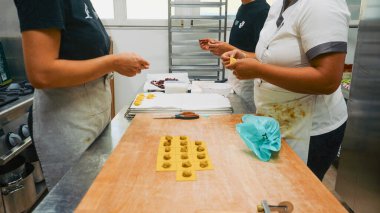 Şefin elleri ahşap bir yüzeyde sarı makarna hamurunu yoğuruyor ve şekillendiriyor. Titiz bir mutfak ortamında el yapımı yemek hazırlama sürecini gözler önüne seriyor.