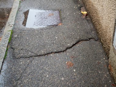 Hasarlı asfalt kaldırım derin bir çatlak ve yıpranmış eski bir metal kapağı gösteriyor.