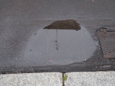 Koyu asfaltta, açık gri beton kaldırım kenarında, sessiz bir su birikintisinde toplanan parlayan yağmur suyu.