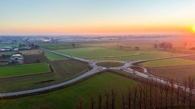 Şehir yollarıyla tarlaları birbirine bağlayan yoğun bir kavşağın havadan görünüşü. Arabalar, dramatik bir günbatımı gökyüzü altında verimli bir ulaşım kavşağından geçiyor.
