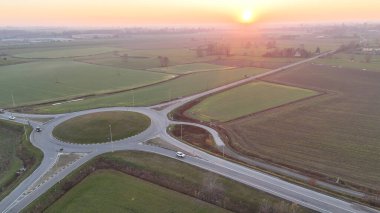 Lombardiya 'nın kırsal kesimlerini birbirine bağlayan modern bir kavşakta trafik dönüyor. Sıcak altın saat ışığı altında yeşil ve kahverengi tarlalarda zikzak çiziyor. Ve huzurlu turuncu bir gün batımı gökyüzü.