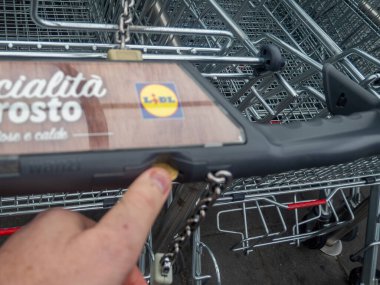 Cremona, İtalya. 25 Ekim 2025. Tüketici Lidl 'de bir alışveriş arabasının kilidini açmak için bozuk para yerleştiriyor. Perakende erişimi, kolaylığı ve indirimleri vurguluyor.