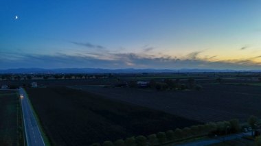 Hava manzarası, engin tarımsal tarlalarda yol alan ıssız bir arabayı yakalıyor. Şehir ışıkları ve uzak dağlar, mavi gökyüzünün altında alacakaranlıkta küçük bir ay görünüyor.