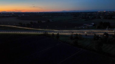 Piacenza Brescia karayolunda gece akan A21 karayolu trafiği, aydınlık bir yol boyunca ilerleyen arabalar ve kamyonlar kentsel ufuk çizgisi ve alacakaranlıkta dağlarla birlikte