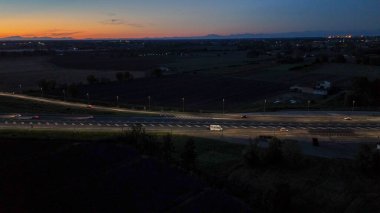 Otoyol trafiği alacakaranlıkta otoyolda ışık ve hareket dinamikleri yaratıyor, turuncu ve mavi gökyüzü altında İtalyan kırsal kesiminde ulaşım ve altyapıyı gösteriyor.