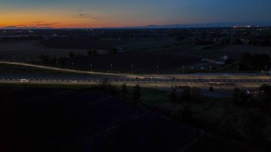 Otoyol trafiği alacakaranlıkta otoyolda ışık ve hareket dinamikleri yaratıyor, turuncu ve mavi gökyüzü altında İtalyan kırsal kesiminde ulaşım ve altyapıyı gösteriyor.