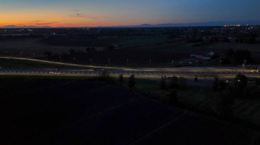 Otoyol trafiği alacakaranlıkta otoyolda ışık ve hareket dinamikleri yaratıyor, turuncu ve mavi gökyüzü altında İtalyan kırsal kesiminde ulaşım ve altyapıyı gösteriyor.