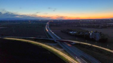 A21 karayolu akşam trafiğini gösteriyor, aydınlık otoyol boyunca ilerleyen araçlar ve üst geçit yolları güzel bir günbatımı gökyüzü altında uzak şehirleri birbirine bağlıyor, modern altyapıyı aydınlatıyor.