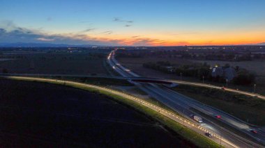 Hava görüntüsü, çok şeritli otoyolda giden araçları ve yolları birleştiren araziyi renkli bir günbatımı gökyüzü altında aydınlatan, bağlantıyı ve yolculuğu sembolize eden manzarayı gösteriyor.