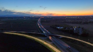 A21 karayolu akşam trafiğini gösteriyor, aydınlık otoyol boyunca ilerleyen araçlar ve üst geçit yolları güzel bir günbatımı gökyüzü altında uzak şehirleri birbirine bağlıyor, modern altyapıyı aydınlatıyor.