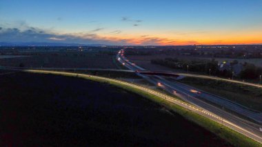 A21 karayolu akşam trafiğini gösteriyor, aydınlık otoyol boyunca ilerleyen araçlar ve üst geçit yolları güzel bir günbatımı gökyüzü altında uzak şehirleri birbirine bağlıyor, modern altyapıyı aydınlatıyor.