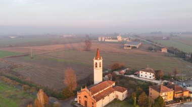 San Bartolomeo kilisesi ve çan kulesi. Vescovato. Krema. İtalya. Puslu bir günbatımında tarımsal tarlalar ve kırsal binalar arasında belirgin bir şekilde duruyor. Lombardiya 'nın sakin kırsalını vurguluyor