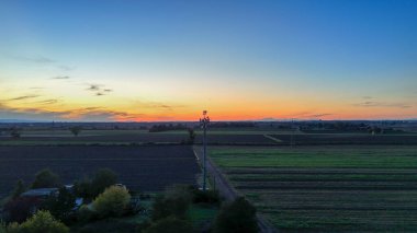 Renkli günbatımı gökyüzüne karşı iletişim baz istasyonu olan Po Vadisi manzarası, İtalyan kırsalındaki bağlantıyı, uzak teknolojiyi ve modern altyapıyı sembolize ediyor.