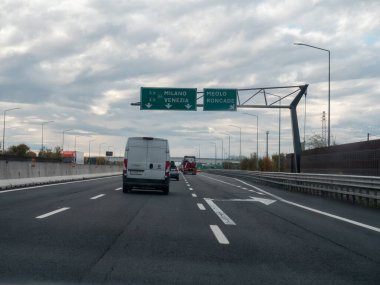 Bulutlu bir günde, yolda beyaz bir minibüs, diğer arabalar ve kamyonları gösteren yönlü işaretlerle otoyolda araçlar sürüyor. Vicenza, İtalya - 21 Kasım 2025
